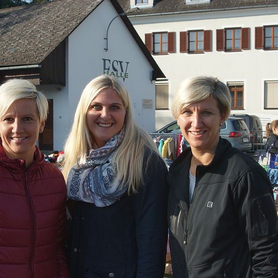 Drei Frauen mit blonden Haaren lächeln vor einem Gebäude mit dem Schild ESV Halle, während ein Auto im Hintergrund geparkt ist.