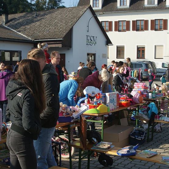 Eine Gruppe von Menschen kauft Spielzeug bei einer Veranstaltung im Freien, mit Tischen voller Artikel vor einem Gebäude mit braunem Rand.