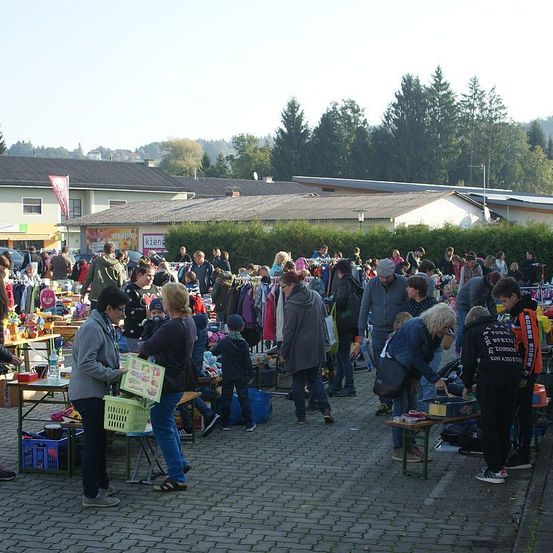 Ein geschäftiger Flohmarkt mit zahlreichen Ständen unter klarem Himmel. Menschen aller Altersgruppen stöbern durch die Artikel. Bäume und Gebäude säumen den Hintergrund.