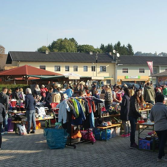 Ein geschäftiger Flohmarkt im Freien mit zahlreichen Ständen, Menschen und ausgestellten Gegenständen unter Zelten und Markisen, vor dem Hintergrund von Gebäuden und Bäumen.