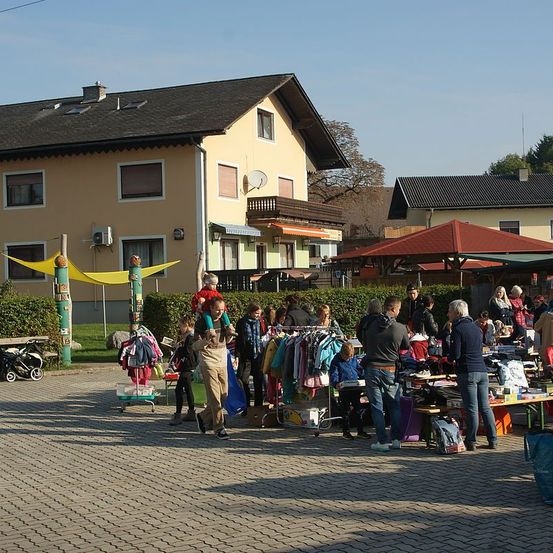 Ein Flohmarkt unter einem roten Baldachin, mit einkaufenden Menschen und spielenden Kindern, vor einem Haus mit Balkon und Satellitenschüssel.