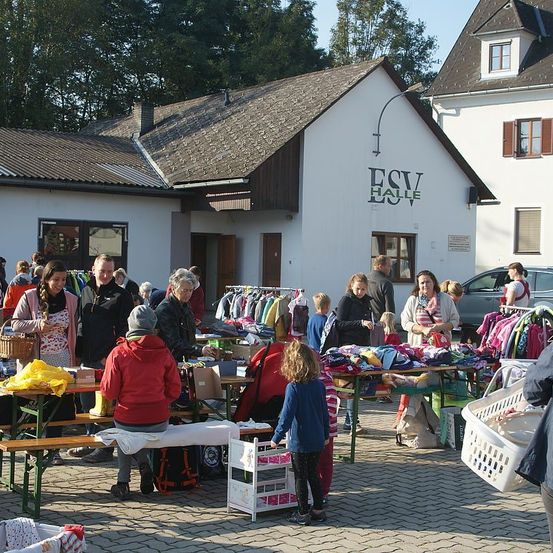 Menschen kaufen bei einem Kleidungsverkauf vor ESV Halle ein. Kleidung ist auf Tischen und Gestellen, und einige Leute sitzen auf Bänken.