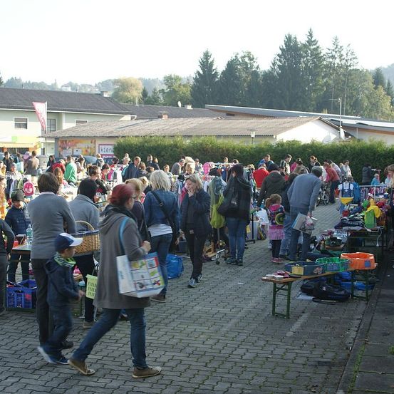 Eine Menschenmenge auf einem Straßenmarkt, die verschiedene Stände mit ausgestellten Artikeln durchstöbert, umgeben von Bäumen und Gebäuden, mit einem klaren Himmel im Hintergrund.