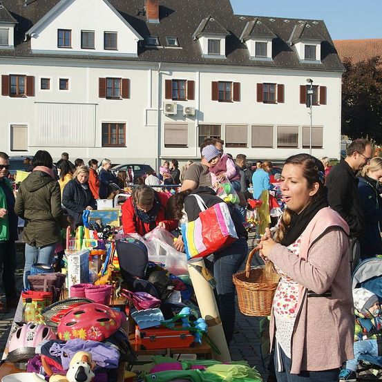 Eine Gruppe von Menschen shoppt auf einem Flohmarkt vor einem weißen Gebäude. Eine Frau schaut sich Spielzeug an und hält einen Korb.