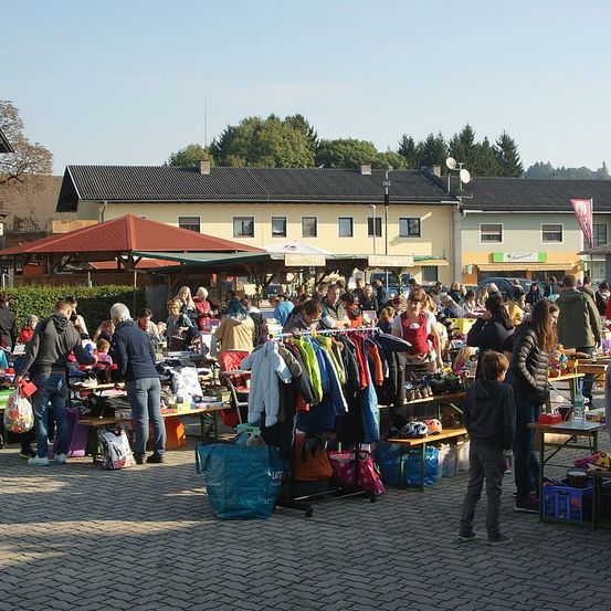 Ein geschäftiger Flohmarkt unter einem roten Baldachin, mit verschiedenen zum Verkauf stehenden Artikeln auf Tischen, umgeben von Menschen und Gebäuden mit Fahnen im Hintergrund.