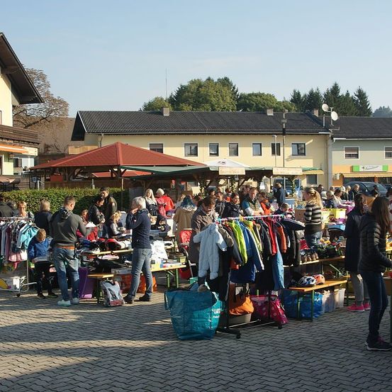 Ein überfüllter Outdoor-Flohmarkt mit zahlreichen Ständen, die verschiedene Artikel verkaufen. Die Stände sind mit bunten Kleidern und Taschen geschmückt. Der Markt ist voller Menschen, einige stöbern und andere kaufen ein. Im Hintergrund bilden Gebäude und Bäume eine malerische Kulisse.