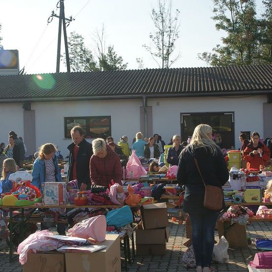 Eine Gruppe von Menschen befindet sich auf einem Flohmarkt vor einem Gebäude. Viele Artikel sind auf den Tischen ausgestellt, darunter Spielzeug und Kisten. Einige Personen stöbern, während andere sich unterhalten.