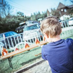 Ein Junge steht draußen in der Nähe eines Zauns und schaut auf ein Spiel mit Flammen darauf. Hinter ihm sind Autos in der Nähe eines Hauses geparkt.