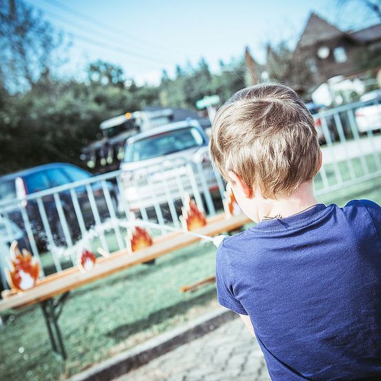 Ein Junge steht draußen in der Nähe eines Zauns und schaut auf ein Spiel mit Flammen darauf. Hinter ihm sind Autos in der Nähe eines Hauses geparkt.