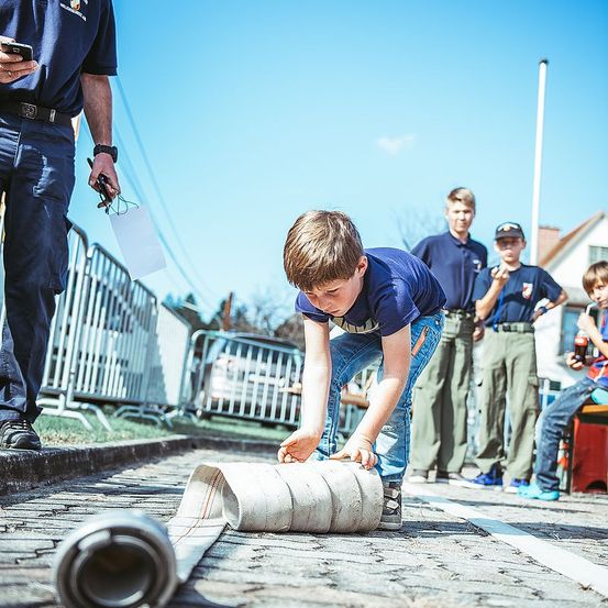 Ein junger Junge beugt sich vor und rollt eine große Schnur auf einem Kopfsteinpflaster, während mehrere Personen hinter ihm stehen.
