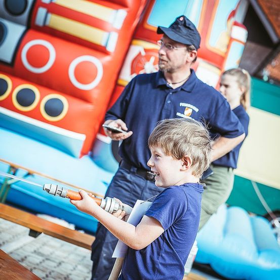 Ein junger Junge hält einen Wasserschlauch und sprüht Wasser, während ein Mann auf sein Handy schaut. Hinter ihnen steht ein buntes aufblasbares Boot mit großen Fenstern.