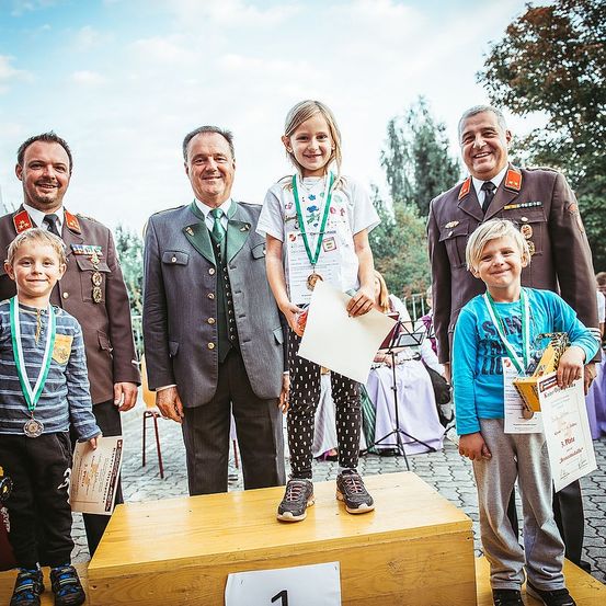 Ein Mädchen auf einem Podest hält eine Urkunde in der Hand, umgeben von Männern in Uniformen, darunter ein Mann mit Medaillen. Hinter ihnen stehen zwei Jungen auf einem weiteren Podest, und ein Mann spielt ein Musikinstrument. Bäume und ein blauer Himmel bilden den Hintergrund.