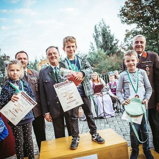 Eine Gruppe von Kindern und Erwachsenen steht auf einem Holzpodium. Die Kinder halten Medaillen und Urkunden. Ein Mann hält einen Blumenstrauß. Dahinter spielt eine Band auf der Bühne.
