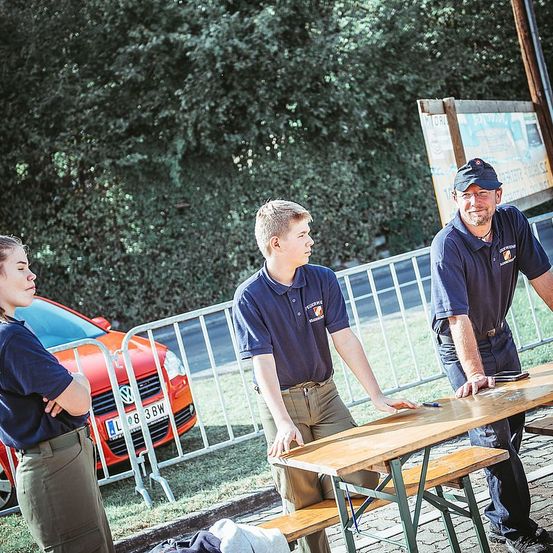 Drei Personen in identischen Uniformen stehen um einen Picknicktisch. Eine Frau schaut nach links, während ein Mann nach unten blickt und der andere Mann lächelt. Ein orangefarbenes Auto ist hinter einem Metallzaun geparkt.