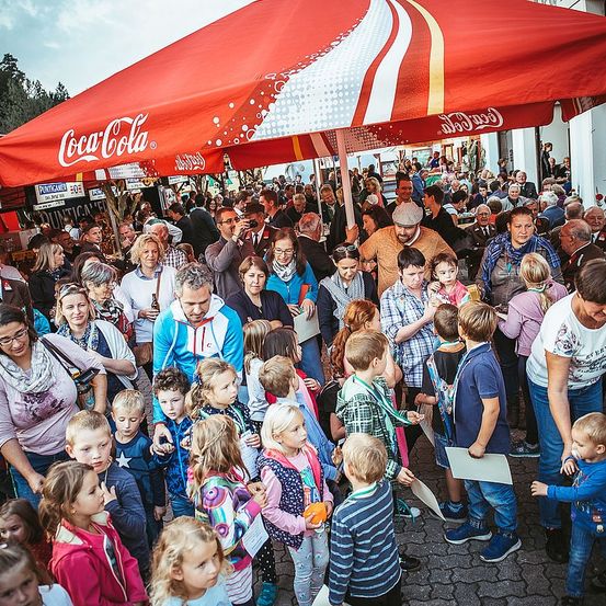 Eine große Menschenmenge versammelt sich unter einem Coca-Cola-Regenschirm. Menschen jeden Alters sind anwesend, darunter Kinder mit Papieren in der Hand, und einige Erwachsene machen Fotos. Die Atmosphäre ist lebhaft, viele Personen führen Gespräche.