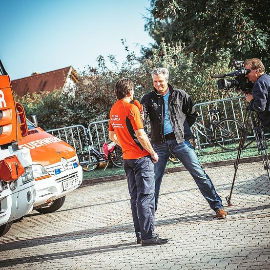 Zwei Männer unterhalten sich in der Nähe eines Krankenwagens und einer Kameracrew. Ein Mann trägt eine blaue Jacke und Jeans, der andere ein orangefarbenes Shirt. Eine Kamera auf einem Stativ ist in der Nähe aufgestellt.
