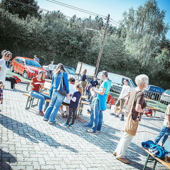 Eine Gruppe von Menschen ist im Freien versammelt, wahrscheinlich auf einem Festival. Einige sitzen, während andere stehen und plaudern und die Atmosphäre genießen. In der Nähe sind Fahrzeuge geparkt, und Bäume bieten eine malerische Kulisse.
