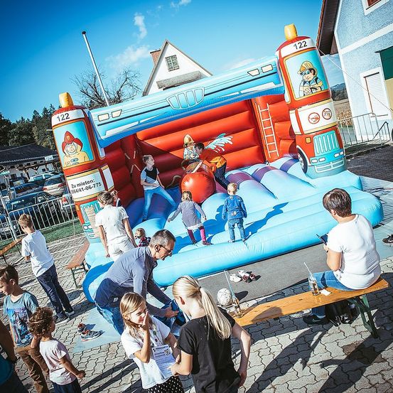 Kinder spielen in einem großen aufblasbaren Feuerwehrauto. Erwachsene beobachten und einige sitzen auf einer Bank. Der Ort ist im Freien, mit Häusern und geparkten Autos im Hintergrund.