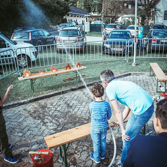 Ein Mann und ein Kind sprühen Wasser aus einem Schlauch auf eine Bank mit brennenden Kerzen. Menschen stehen herum und Autos sind in der Nähe geparkt.