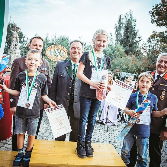 Eine Gruppe von Kindern und Erwachsenen steht auf einer Holzplattform, lächelt und hält Medaillen und Urkunden in den Händen. Die Kinder tragen Medaillen und halten Urkunden, während die Erwachsenen hinter ihnen stehen. Im Hintergrund sind Bäume und eine Statue zu sehen.