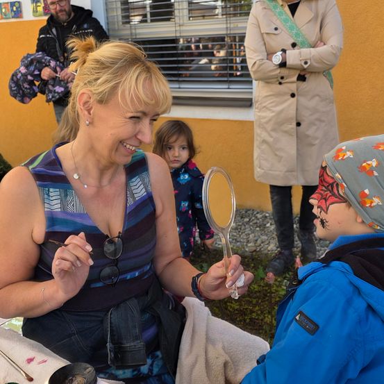 Eine Frau malt ein Spinnennetz auf das Gesicht eines Kindes. Dahinter beobachten ein Mädchen und ein erwachsener Mann. Eine weitere Frau steht rechts und beobachtet. Die Wand im Hintergrund ist gelb.