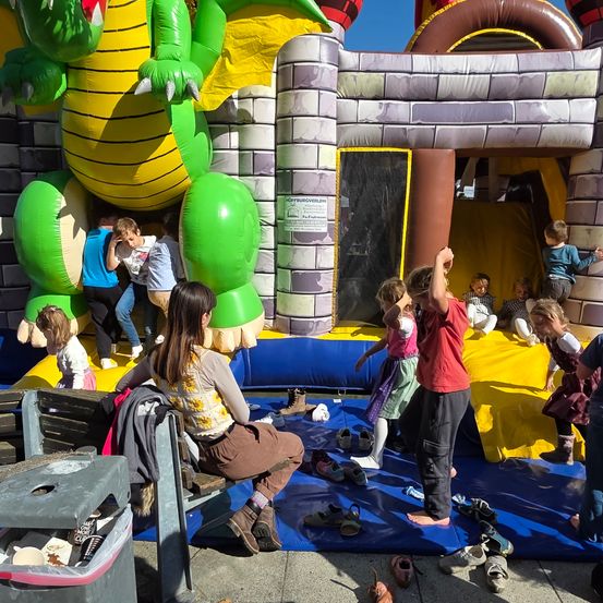 Kinder spielen in einem großen aufblasbaren Schloss, während andere ihre Schuhe auf einem blauen Teppich ausziehen. Einige klettern auf das Aufblasbare, andere sitzen auf dem Boden.