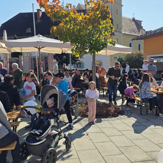 Im Freien gelegener Essbereich mit Tischen und Stühlen unter Sonnenschirmen. Menschen, darunter auch Kinder, sitzen und stehen herum. Ein Baby ist in einem Kinderwagen in der Nähe eines Baumes.
