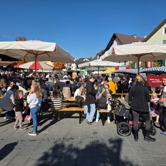 Ein überfüllter Außenbereich mit vielen Menschen, die unter weißen Sonnenschirmen an einem sonnigen Tag sitzen. Einige unterhalten sich, während andere in ihre Handys schauen. Der Bereich ist voller Aktivität.