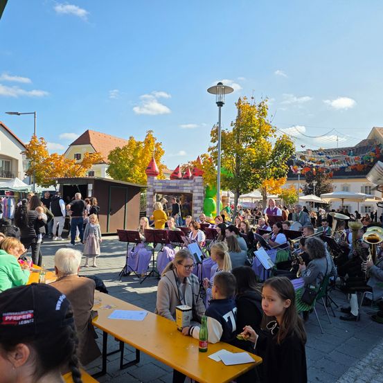Eine Gruppe von Menschen, darunter Kinder, versammelt sich an einem Außenort, möglicherweise für ein Festival. Einige sitzen um Tische herum, andere stehen. Musikinstrumente und eine kleine Bühne mit einem aufblasbaren Schloss sind vorhanden. Das Gelände ist von Bäumen umgeben, und Gebäude sind im Hintergrund zu sehen.