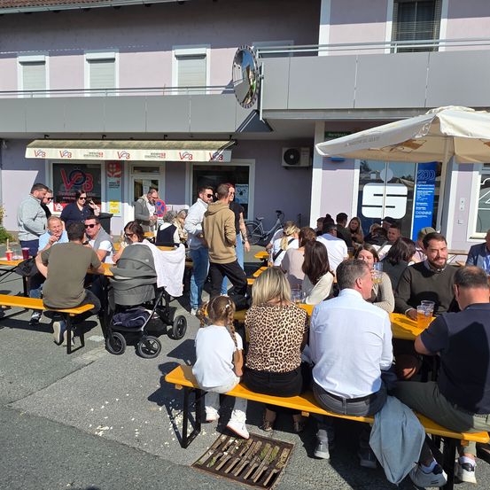 Eine Gruppe von Menschen sitzt an Tischen vor einem Gebäude mit Markisen und einem Regenschirm. Einige tragen Sonnenbrillen und andere haben Getränke dabei. In der Nähe befinden sich ein Kinderwagen und Fahrräder.