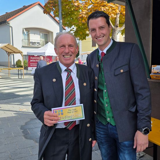 Zwei Männer lächeln und posieren für ein Foto. Ein Mann hält ein gelbes Ticket und trägt eine rot-weiß gestreifte Krawatte. Der andere Mann trägt ein grünes Weste und blaue Jeans. Hinter ihnen ist ein unscharfer Hintergrund mit einer gelben Bank und einem Baum.