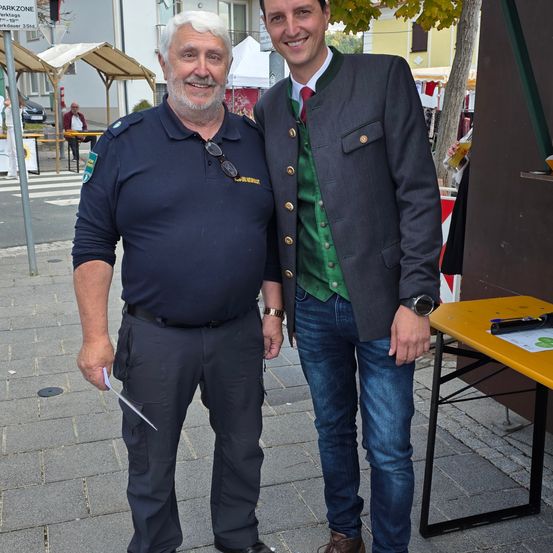 Zwei Männer stehen auf einem Bürgersteig, einer hält ein Stück Papier und lächelt in die Kamera. Ein Tisch und ein Baum sind im Hintergrund.