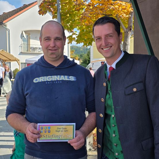 Zwei Männer stehen eng beieinander und lächeln in die Kamera. Ein Mann hält eine gelbe Karte.