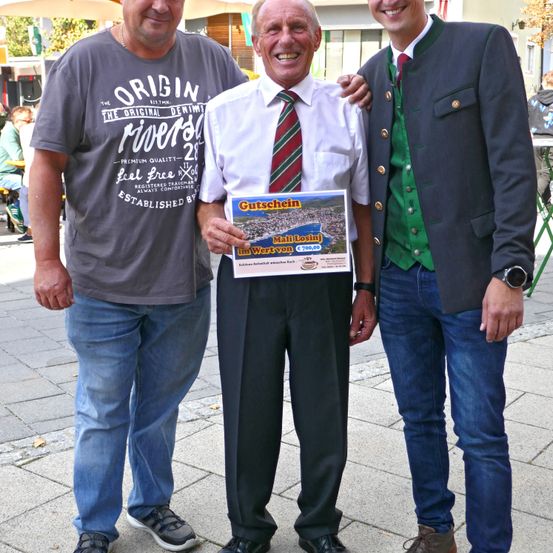 Drei Männer stehen draußen und lächeln. Der Mann in der Mitte hält eine Broschüre mit einer Karte von Gutschien. Der Mann links trägt ein graues Origin River T-Shirt. Der Mann rechts trägt einen dunklen Anzug.