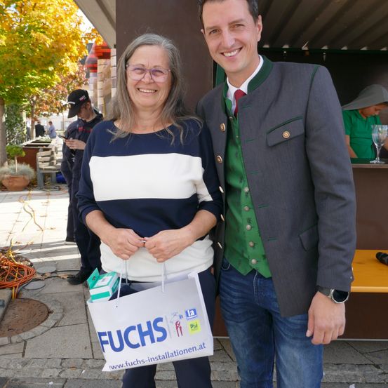 Eine lächelnde ältere Frau und ein Mann in traditioneller deutscher Kleidung stehen draußen und halten einen Einkaufsbeutel mit der Aufschrift 'FUCHS'. Hinter ihnen stehen Leute und benutzen ihre Handys.
