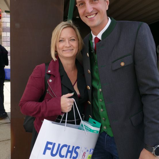 Ein lächelnder Mann und eine Frau stehen zusammen, der Mann in einer grünen Weste und die Frau hält einen Fuchs-Einkaufstasche. Sie scheinen für ein Foto zu posieren.