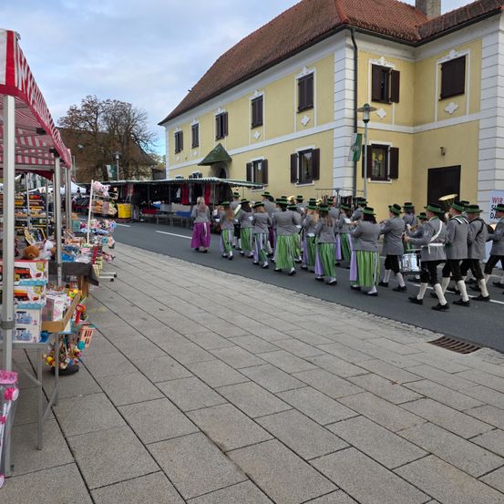 Eine Blaskapelle spielt Instrumente, während sie in einer Reihe vor einem Gebäude mit Geschäften und Ständen in der Nähe geht.