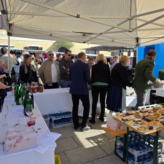 Eine Gruppe von Menschen versammelt sich unter einem Zelt bei einer Outdoor-Veranstaltung und betrachtet Essen und Wein auf den Tischen.