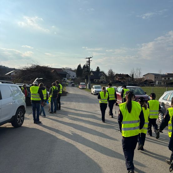 Eine Gruppe von Menschen in gelben Westen geht eine Straße entlang. Autos sind auf beiden Seiten der Straße geparkt. Der Himmel ist blau mit einigen Wolken.