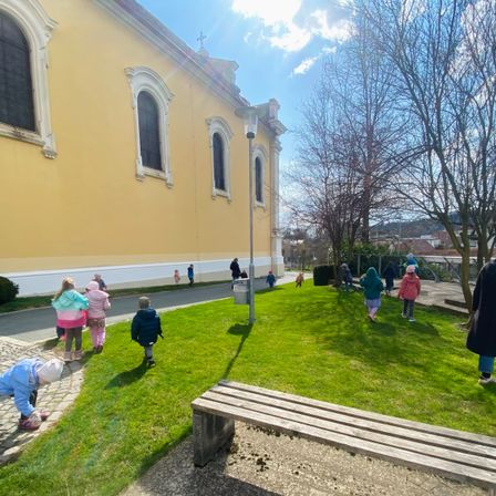 Kinder spielen vor einer Kirche an einem sonnigen Tag. Im Vordergrund steht eine Bank.