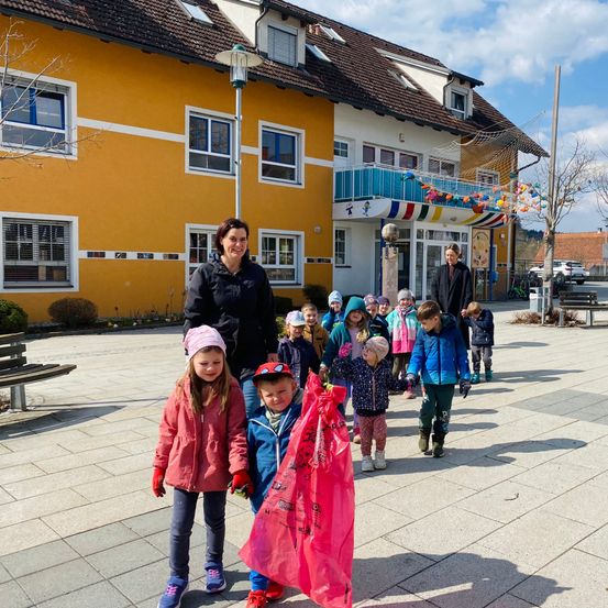 Eine Gruppe von Kindern und zwei Erwachsenen geht vor einem gelben Gebäude. Die Kinder tragen Winterkleidung, und einer hält eine rosa Tasche. Die Erwachsenen lächeln.
