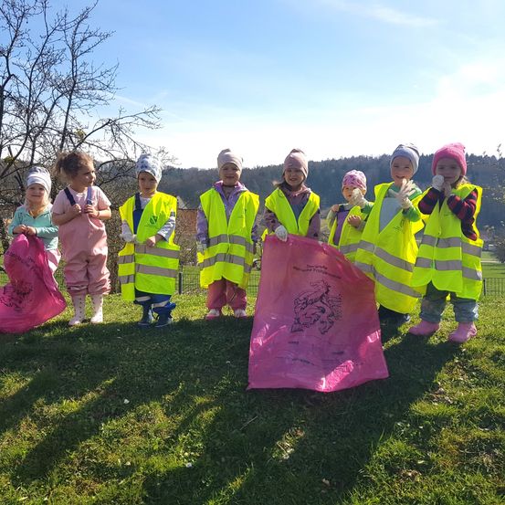 Eine Gruppe von Kindern in hellgelben Westen und Handschuhen steht auf einem Grasfeld und hält rosa Tüten. Sie lächeln und scheinen an einer Aufräumaktion teilzunehmen. Dahinter steht ein blattloser Baum und ein klarer blauer Himmel.