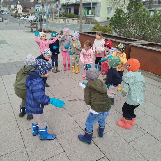 Eine Gruppe von Kindern in Winterkleidung steht draußen im Kreis, einige mit Handschuhen, andere mit Mützen. Hinter ihnen gibt es Pflanzen, ein Gebäude und eine Straße mit geparkten Autos.
