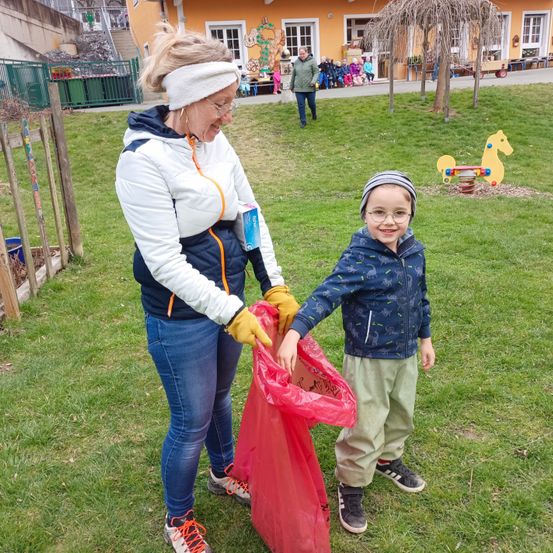 Eine Frau und ein Junge halten einen roten Müllsack und stehen auf einem grünen Rasen. Dahinter sind ein Gebäude, ein Pferdespielzeug und eine Gruppe von Kindern zu sehen.
