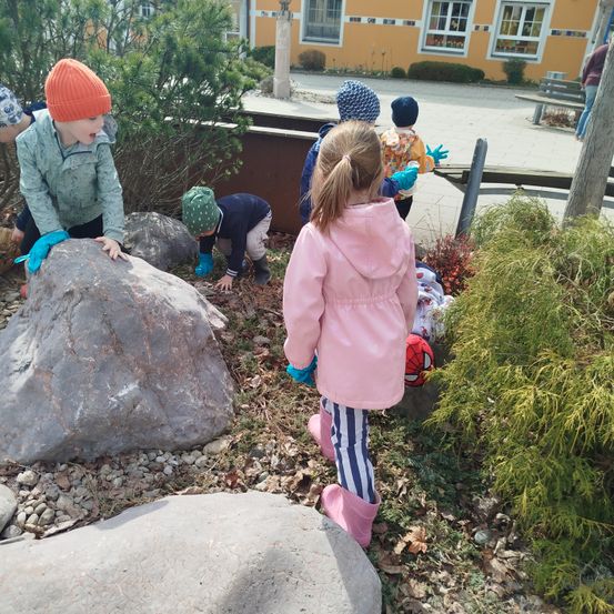 Kinder in warmen Kleidern erkunden einen felsigen Bereich mit Pflanzen. Ein Kind trägt eine pinke Mütze und rosa Stiefel. Hinter ihnen ein Gebäude mit gelben Wänden.