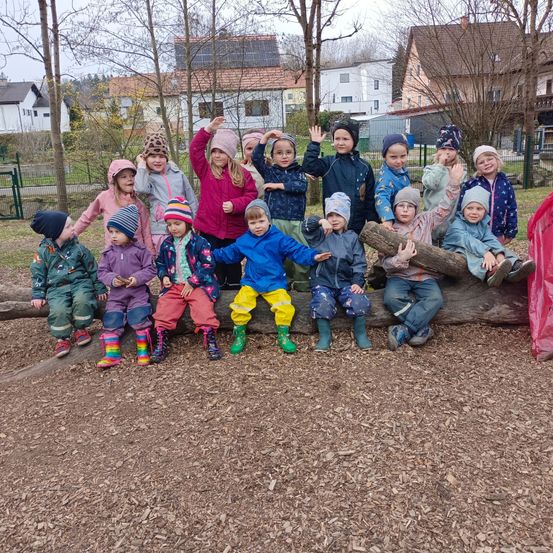 Eine Gruppe junger Kinder in Winterkleidung posiert für ein Foto auf einem Baumstamm in einem Park, mit Häusern und Bäumen im Hintergrund.