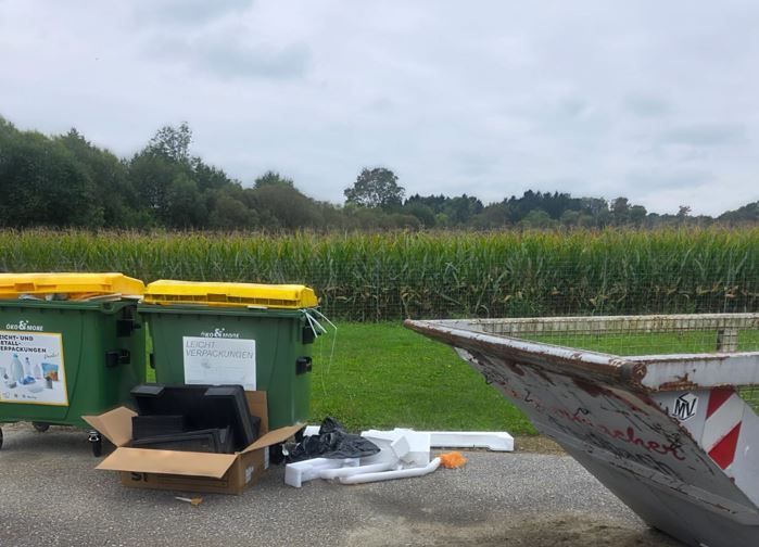 Zwei grüne Recyclingbehälter und ein Pappkarton mit Elektronik befinden sich in einem Feld mit Maisfeld und Zaun im Hintergrund. Ein verrosteter Metallcontainer ist ebenfalls zu sehen.
