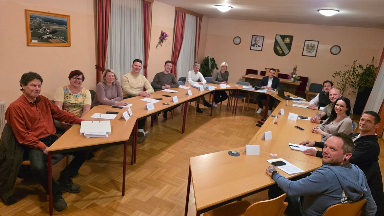 Sieben Personen sitzen in einem Besprechungsraum um einen Tisch herum und unterhalten sich. Papiere, Stifte und ein Laptop liegen auf dem Tisch. Zwei Bilderrahmen hängen an der Wand.