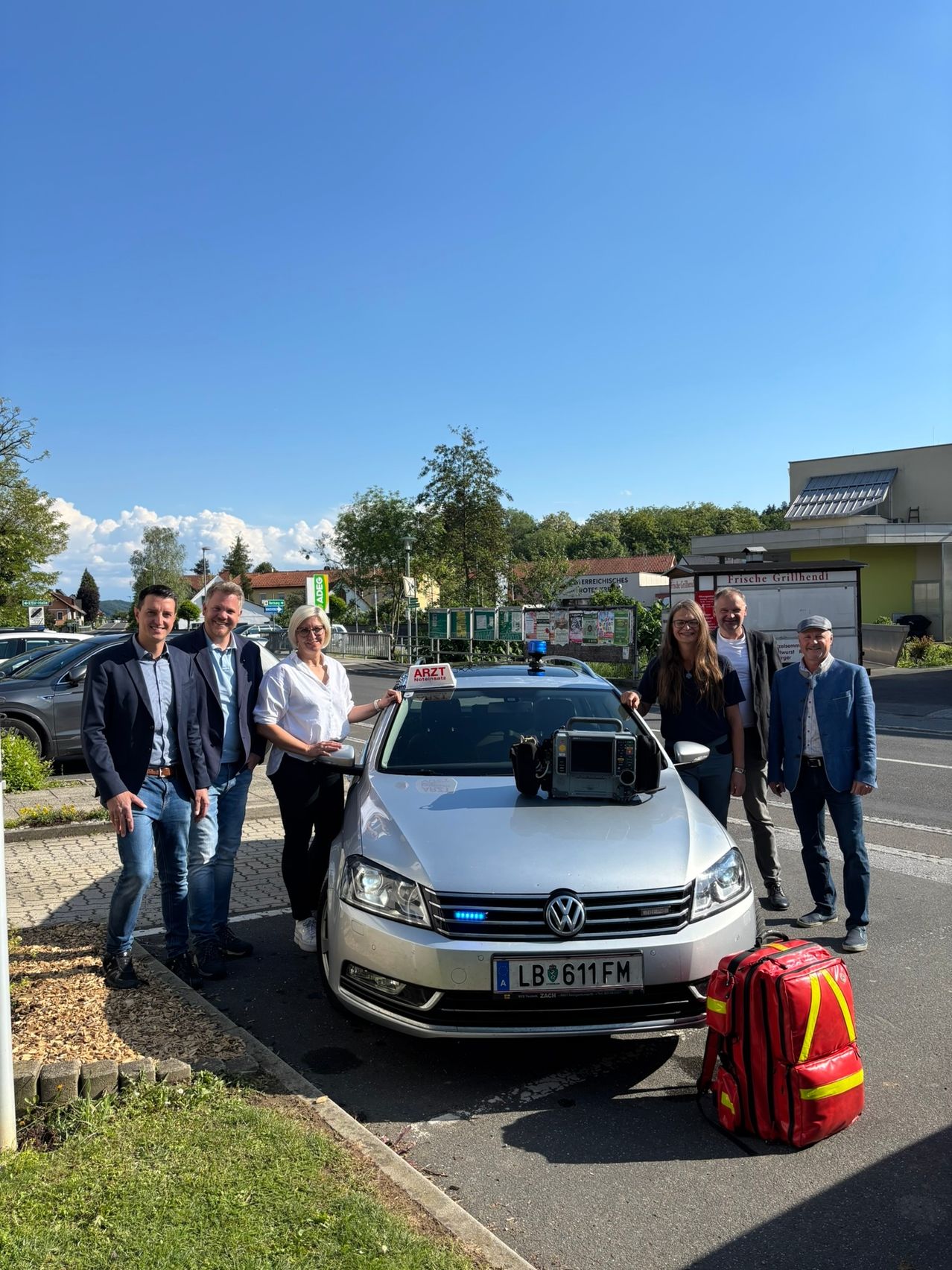 Sechs Personen stehen neben einem silbernen Volkswagen Passat mit einer medizinischen Ausrüstungsbox auf dem Dach. Das Auto hat das Kennzeichen LB 611 FM. Ein roter Rucksack mit gelben und schwarzen Streifen steht neben ihnen. Bäume, ein Gebäude und geparkte Autos sind im Hintergrund.