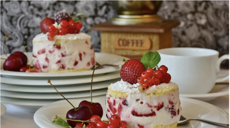 Zwei Desserts mit Erdbeeren und roten Beeren auf weißen Tellern. Ein Kaffeemühle und eine weiße Kaffeetasse im Hintergrund.